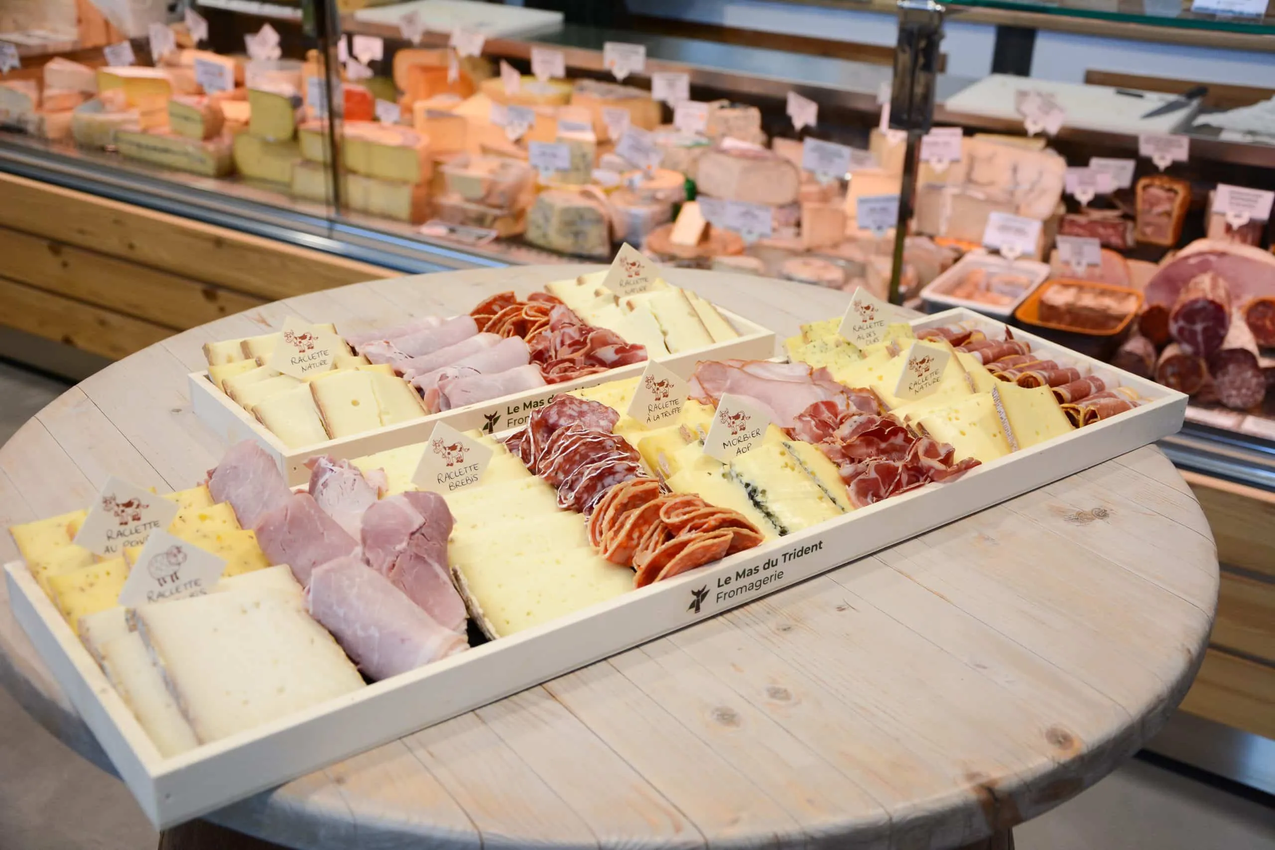 Grand plateau de raclette et de charcuterie posé devant la vitrine à fromages de la boutique