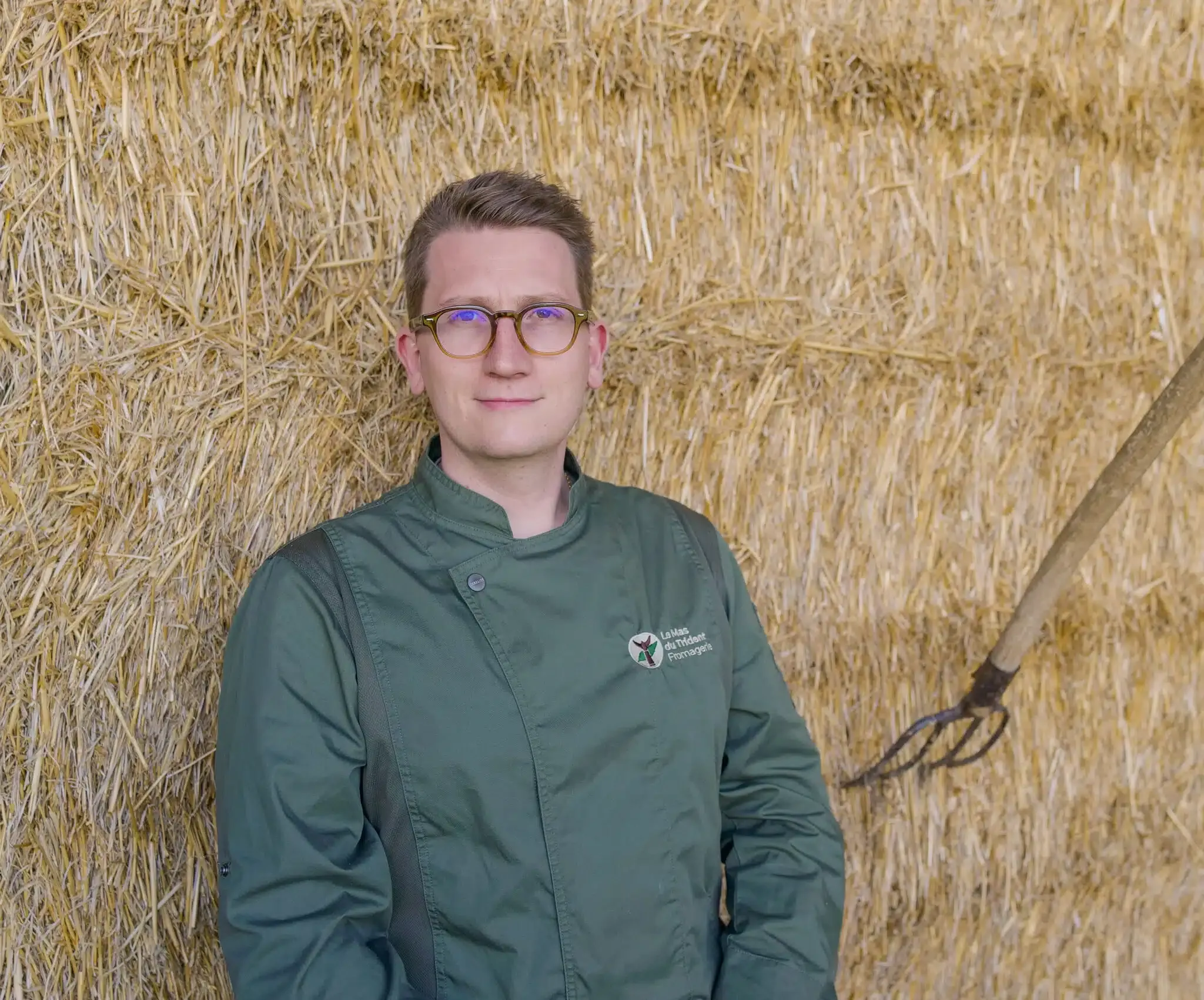 Louis, crémier-fromager, en veste de travail devant une botte de paille
