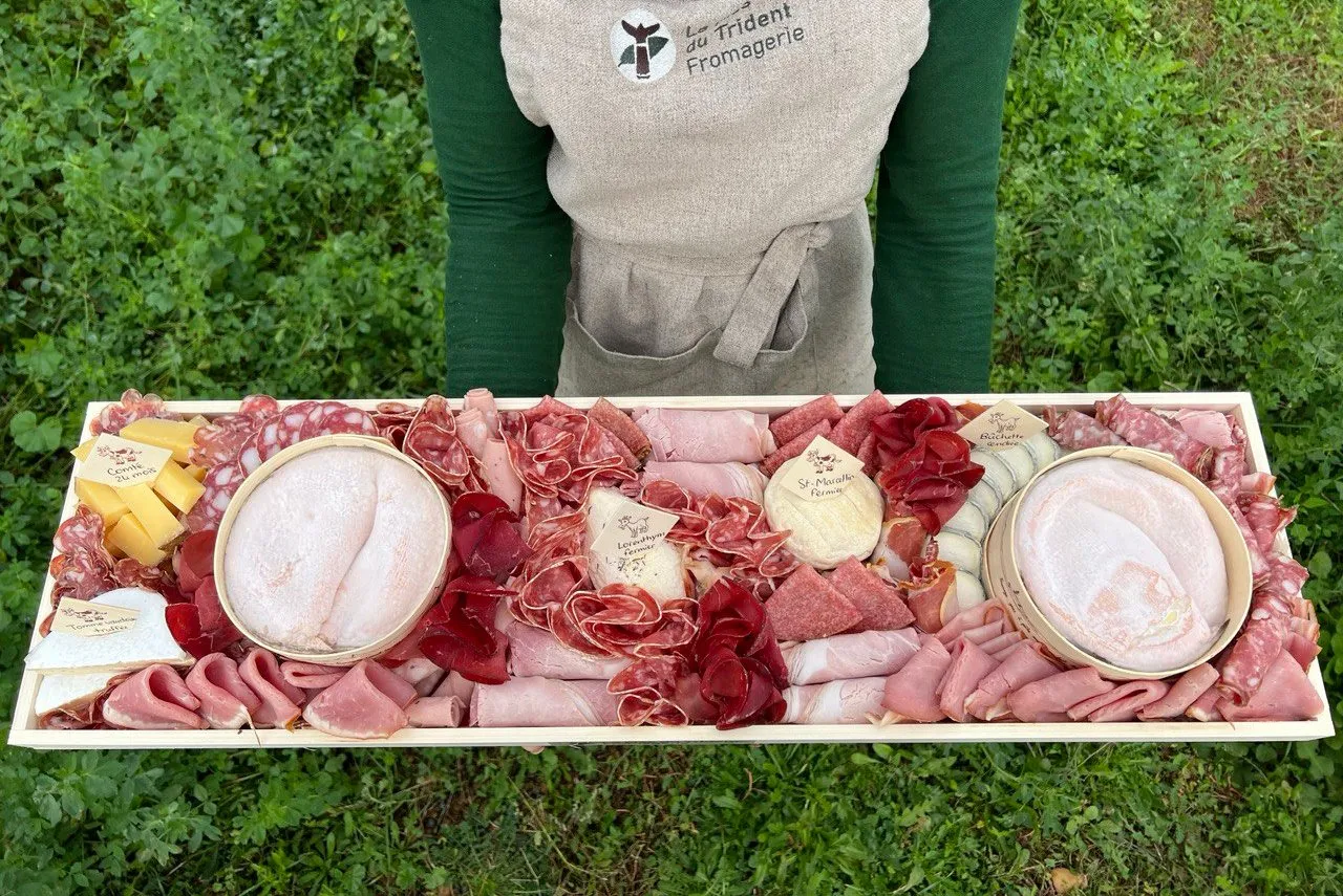 Un grand plateau de charcuterie et de fromages étiquetés, présenté à deux mains par une personne en tablier du Mas du Trident, dehors dans l’herbe