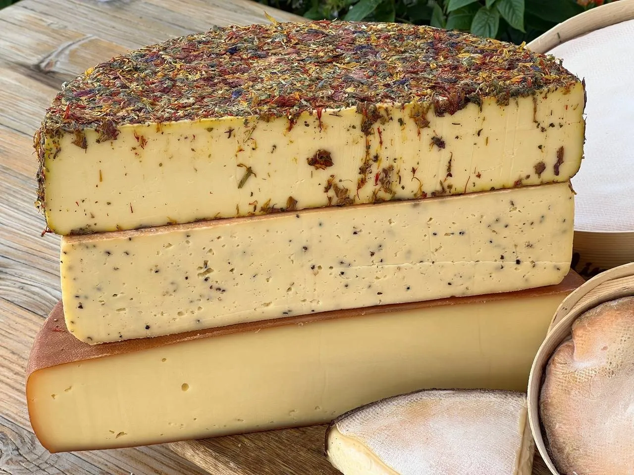 Trois meules de raclette empilées et entamées : l’une à croûte de fleurs séchées, l’autre parsemée de grains de poivre, la troisième nature à croûte lavée